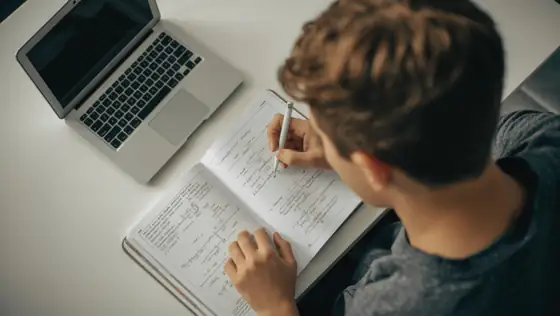 High school student writing math notes in a notebook at a desk with a laptop.